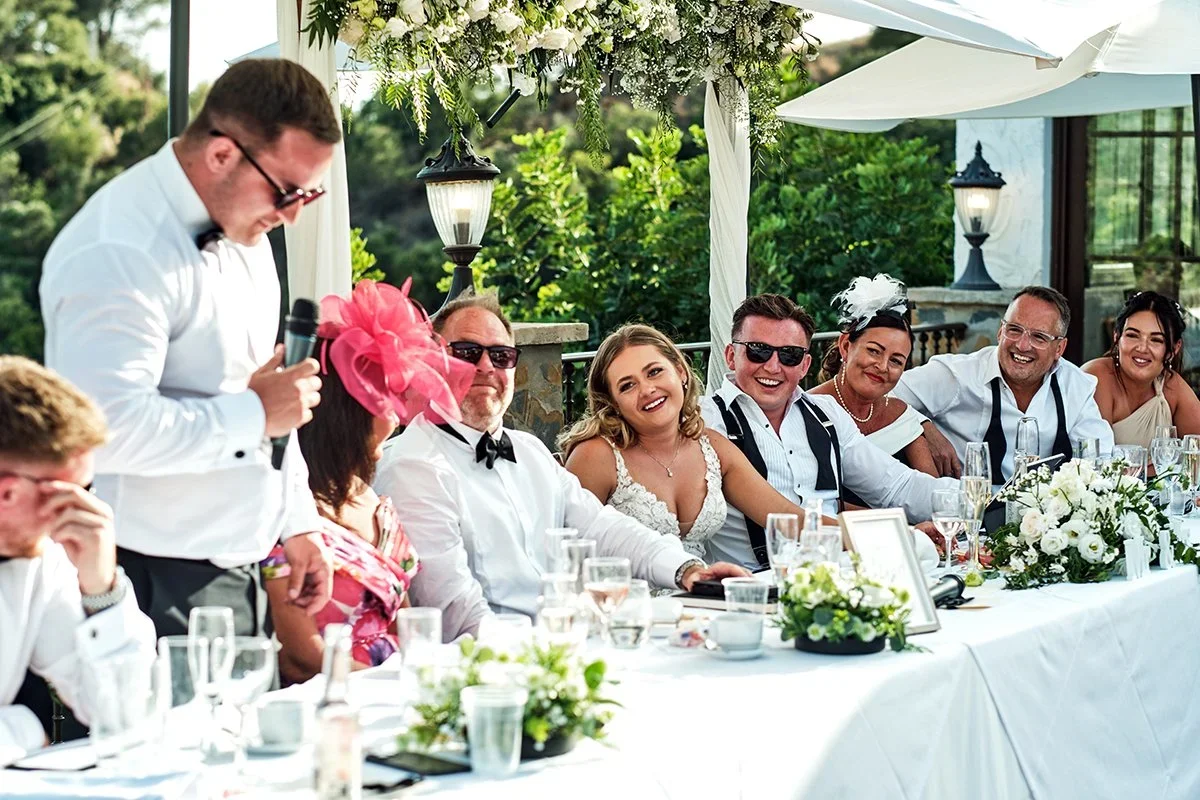 Mesa presidencial en una terraza al aire libre durante un banquete de boda: un hombre de pie con micrófono da un discurso mientras la novia y otros invitados, algunos con gafas de sol y tocados, ríen a carcajadas bajo un arco floral.