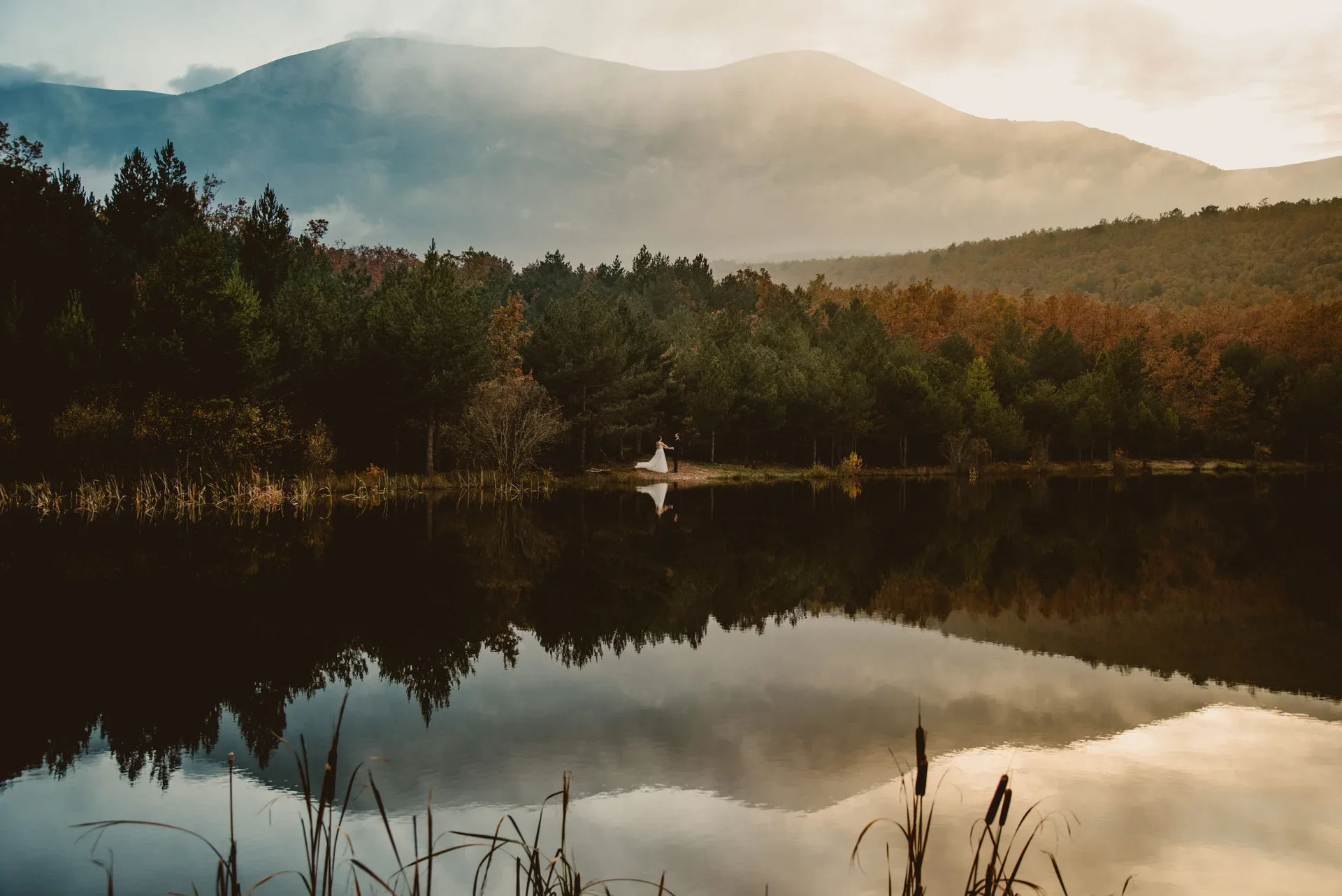 Paisaje de montaña con niebla y un lago en calma que refleja perfectamente el bosque otoñal y las cumbres, con la pareja de novios como pequeñas figuras junto a la orilla.
