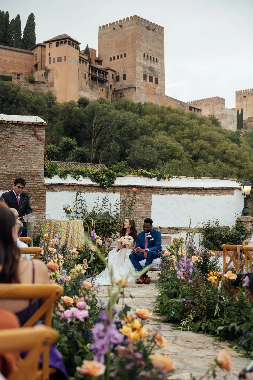 Ceremonia de boda al aire libre con los novios sentados durante el discurso del oficiante, en una terraza con muros de piedra rodeada de flores de colores, con la Alhambra de Granada al fondo sobre la colina.