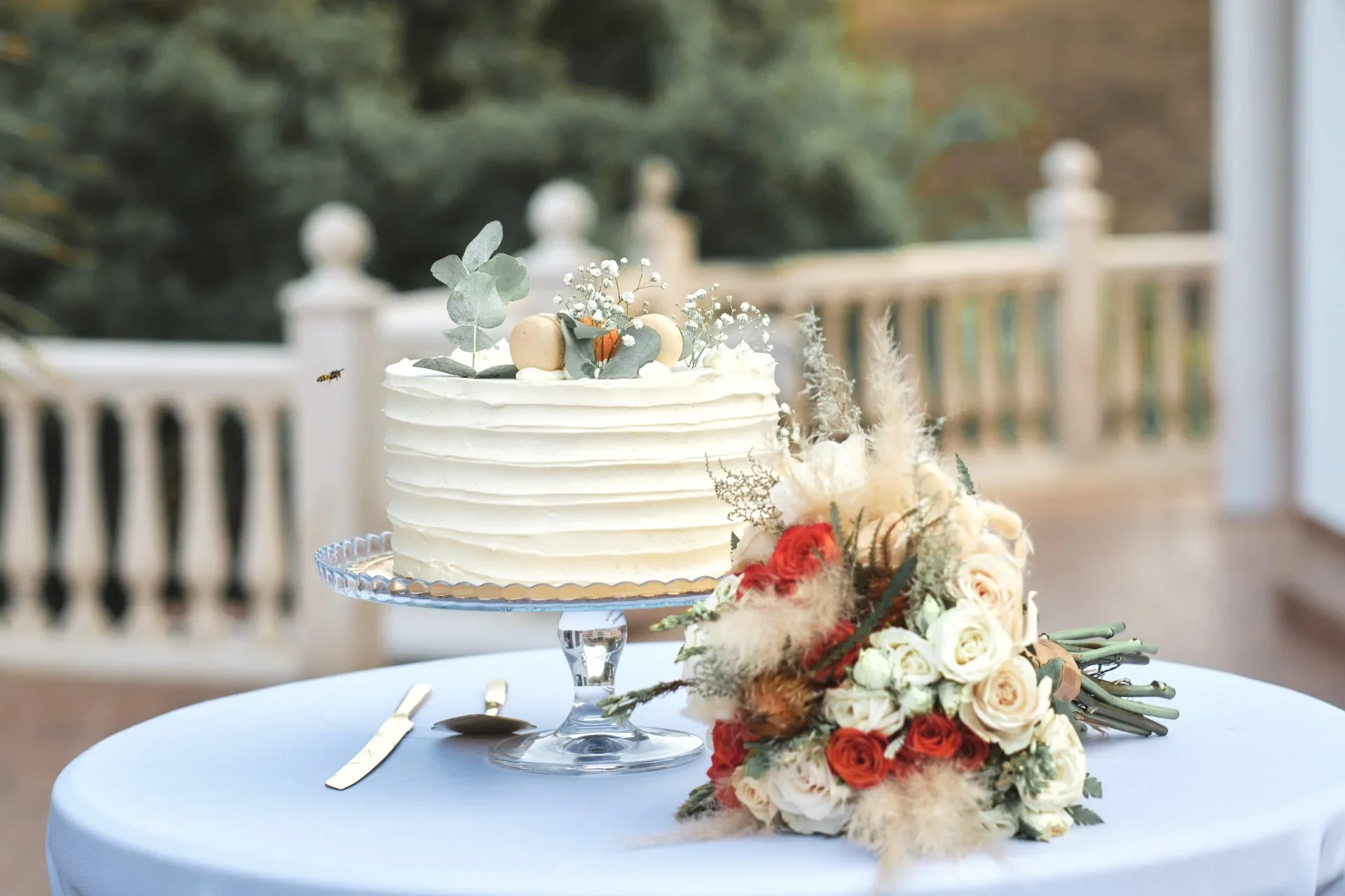 Detalle de una tarta nupcial de un piso decorada con macarons, eucalipto y flores blancas junto a un ramo de novia de rosas rojas y blancas con pampas, sobre una mesa con mantel azul claro y balaustrada desenfocada al fondo.