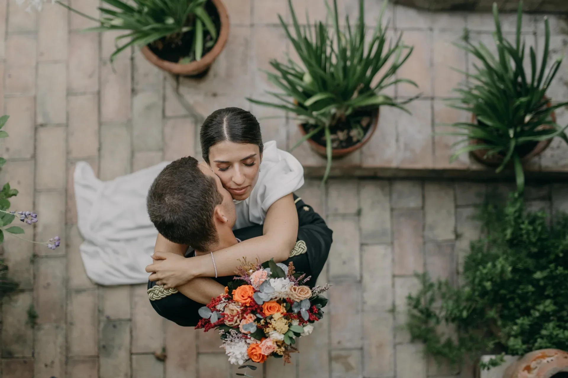 Vista cenital de la pareja abrazándose en un patio de suelo de baldosa terracota; ella, con vestido blanco, apoya la cara sobre el hombro de él, que viste un uniforme militar de gala, mientras sostiene un ramo de rosas naranjas y flores blancas rodeado de macetas con plantas verdes.
