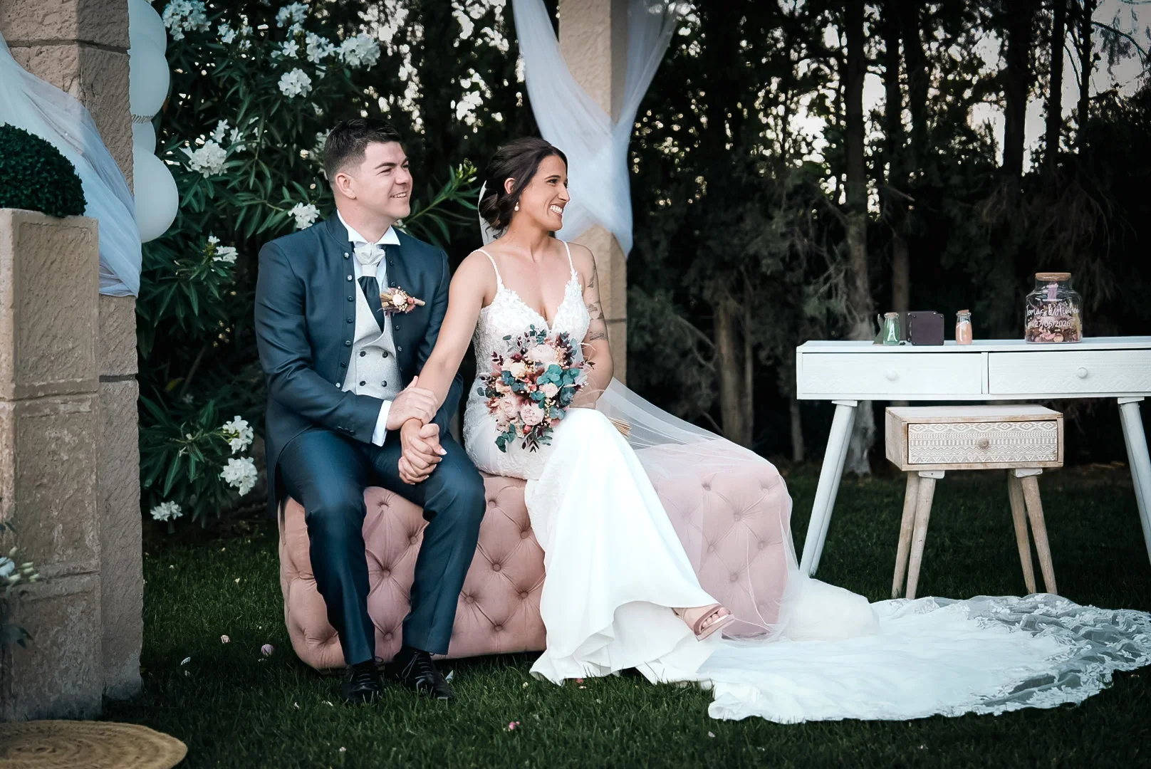 Los novios sentados juntos y sonrientes en un banco tapizado rosa bajo un cenador de piedra con telas blancas, cogidos de la mano, con un jardín de pinos al fondo en la hora dorada.