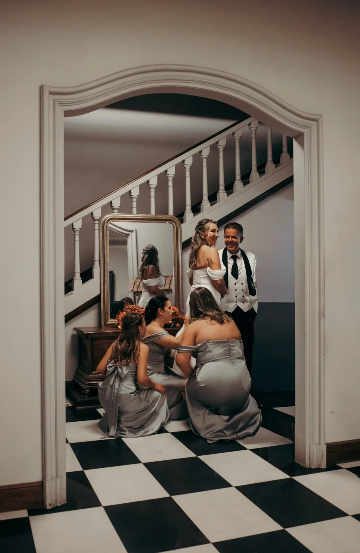 Escena de preparativos captada a través de un arco de puerta: la novia sonríe junto a su padre mientras tres damas de honor con vestidos grises arrodilladas ajustan la cola del vestido, con escalera y espejo dorado de fondo sobre suelo ajedrezado.