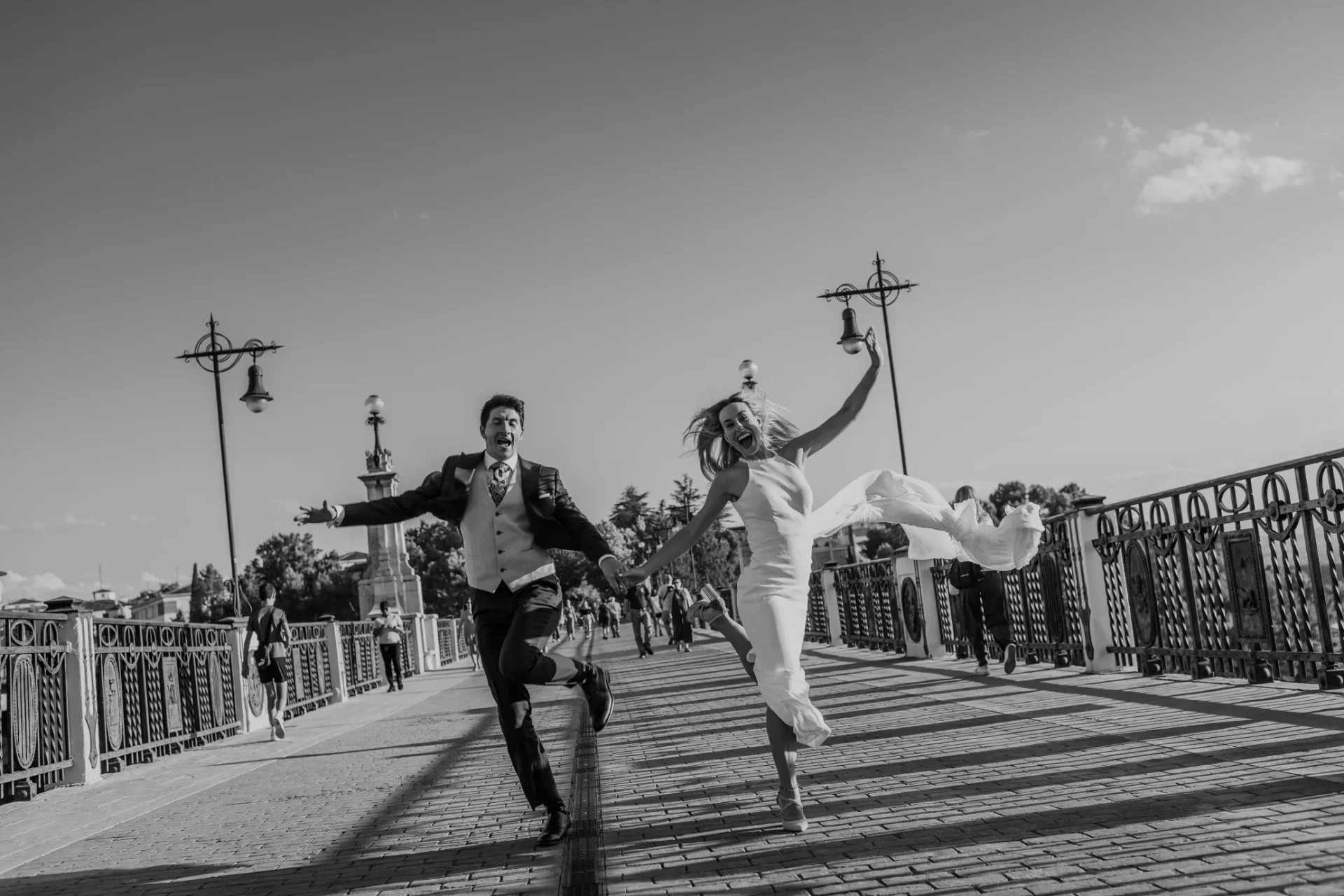 Pareja de recién casados corre saltando de la mano sobre un puente empedrado, en blanco y negro, con farolas antiguas de hierro forjado y transeúntes al fondo; el vestido de ella vuela con el movimiento.