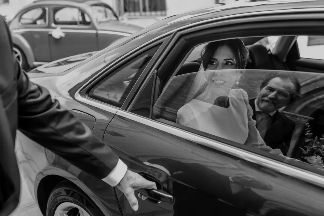 En blanco y negro, la novia sonríe desde el asiento trasero de un coche clásico mientras un hombre le abre la puerta desde fuera, con el reflejo de otro coche de época visible al fondo de la calle.