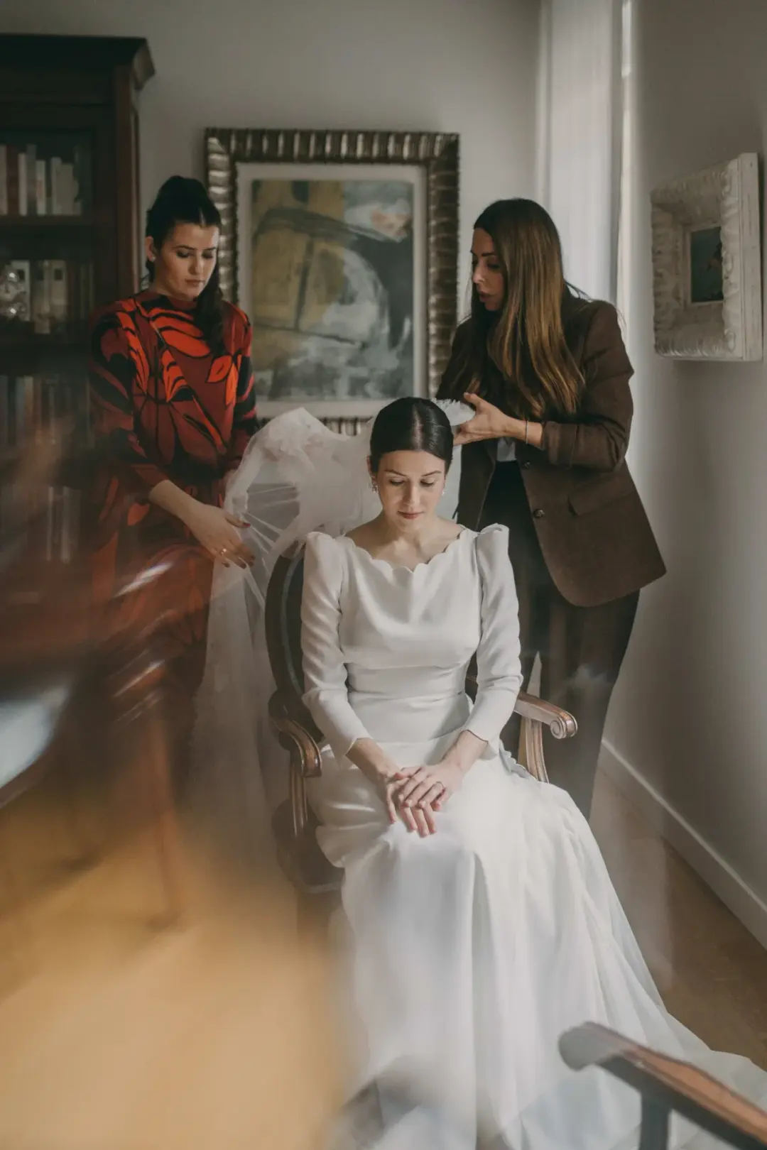 Sentada en una silla antigua, la novia con vestido de cuello festoneado recibe los últimos retoques del velo por parte de dos mujeres, una con vestido rojo estampado y otra con americana marrón, en una estancia con estanterías de libros y un cuadro abstracto enmarcado.