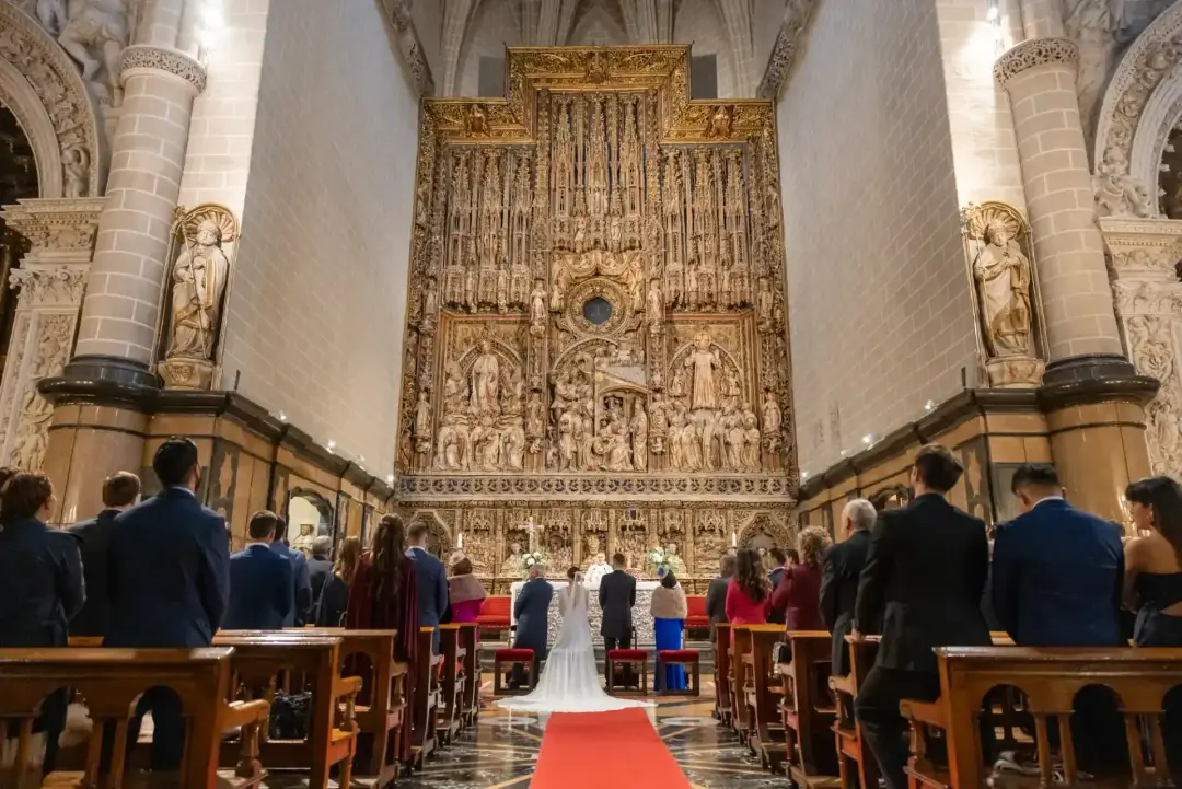 Vista general del interior de una iglesia gótica durante la ceremonia, con los novios ante un retablo dorado monumental, alfombra roja en el pasillo central y los invitados de pie en los bancos.