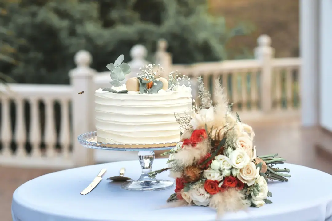 Detalle de una tarta nupcial de un piso decorada con macarons, ramitas de eucalipto y flores blancas junto a un ramo de novia de rosas rojas y blancas con pampas, sobre una mesa con mantel azul claro y balaustrada desenfocada al fondo.