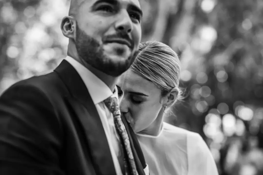Fotografía monocroma de una pareja entrelazada en un abrazo. Ella cierra los ojos y descansa la mejilla contra el cuello y la corbata de él, que dirige la mirada hacia un costado con expresión tranquila. Detrás, unas plantas fuera de foco se disuelven en manchas de luz brillante.