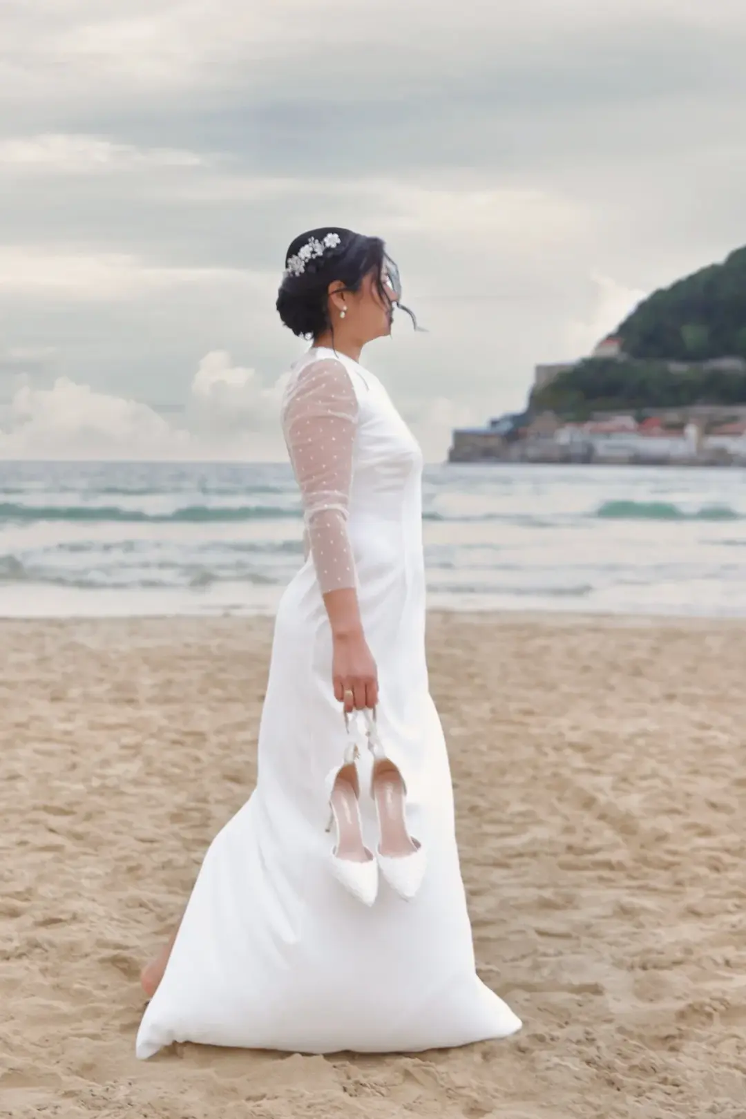 La novia posa de perfil en la playa sujetando sus zapatos de tacón en la mano, con el vestido ondeando al viento y una localidad costera con acantilado al fondo.