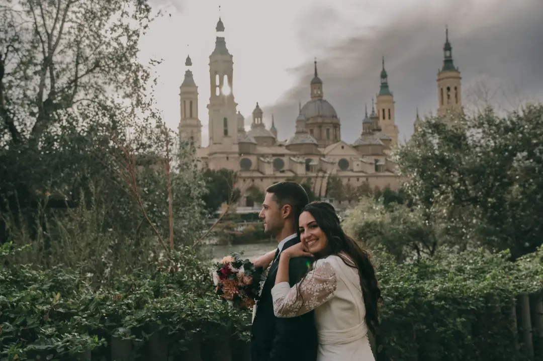 Pareja de novios abrazada, ella sonriendo hacia la cámara con un ramo de flores en tonos coral y burdeos, frente a la Basílica del Pilar de Zaragoza bajo un cielo nublado y dramático, rodeados de vegetación.