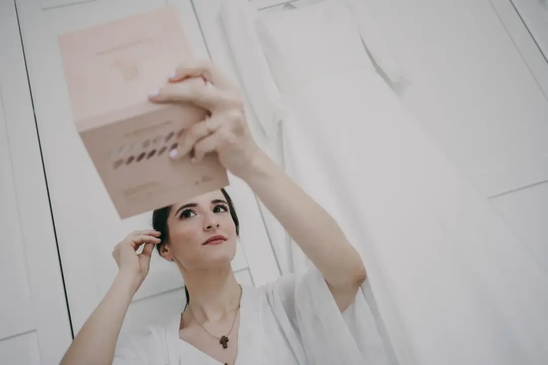 La novia, en bata blanca, sostiene sobre su cabeza una caja rosa de invitaciones o regalo y la observa con atención, en una habitación luminosa y sobreexpuesta con el vestido de novia colgado desenfocado al fondo.
