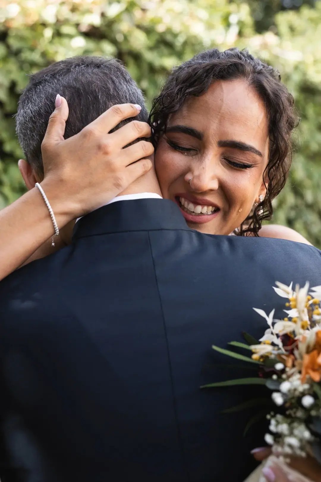 Primer plano emotivo de una invitada llorando y sonriendo mientras abraza a un hombre mayor de espaldas, con un ramo de flores silvestres visible en primer término y follaje verde desenfocado detrás.