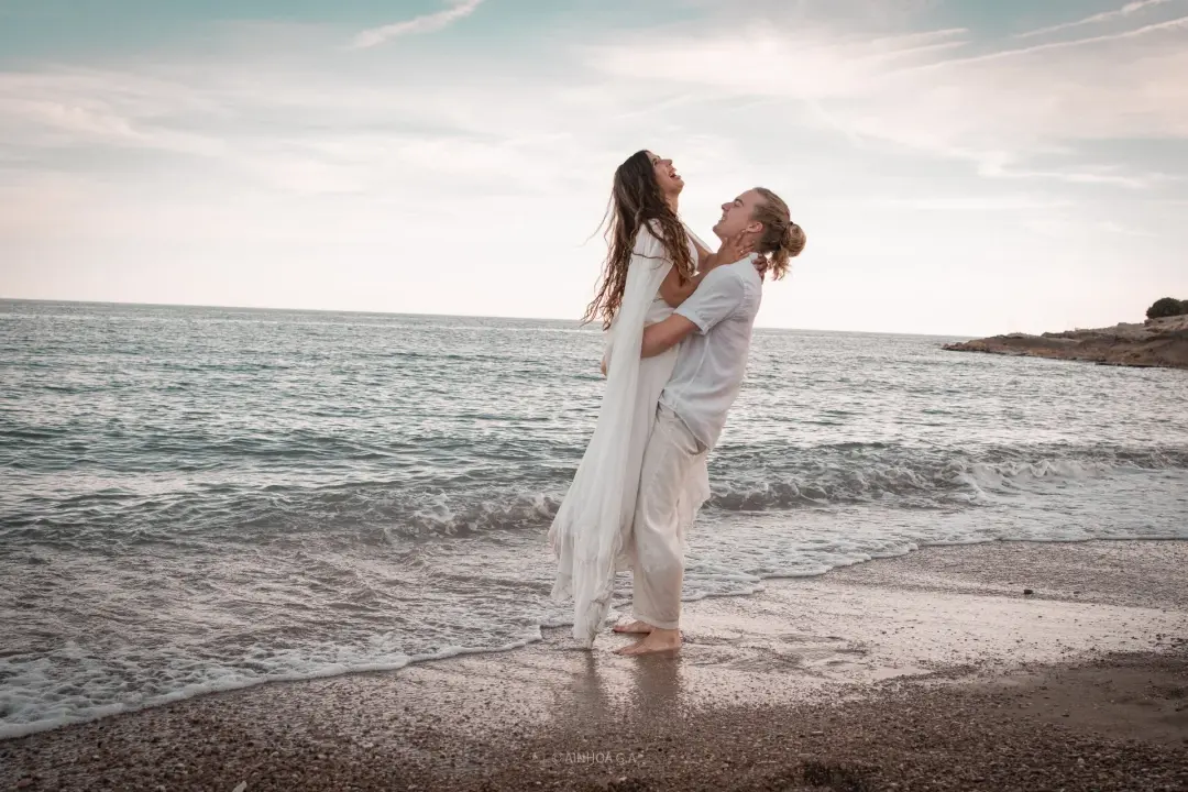 El chico sostiene en volandas a su pareja entre la orilla del mar, ambos riendo mirándose hacia arriba, con el mar y el cielo del atardecer de fondo.