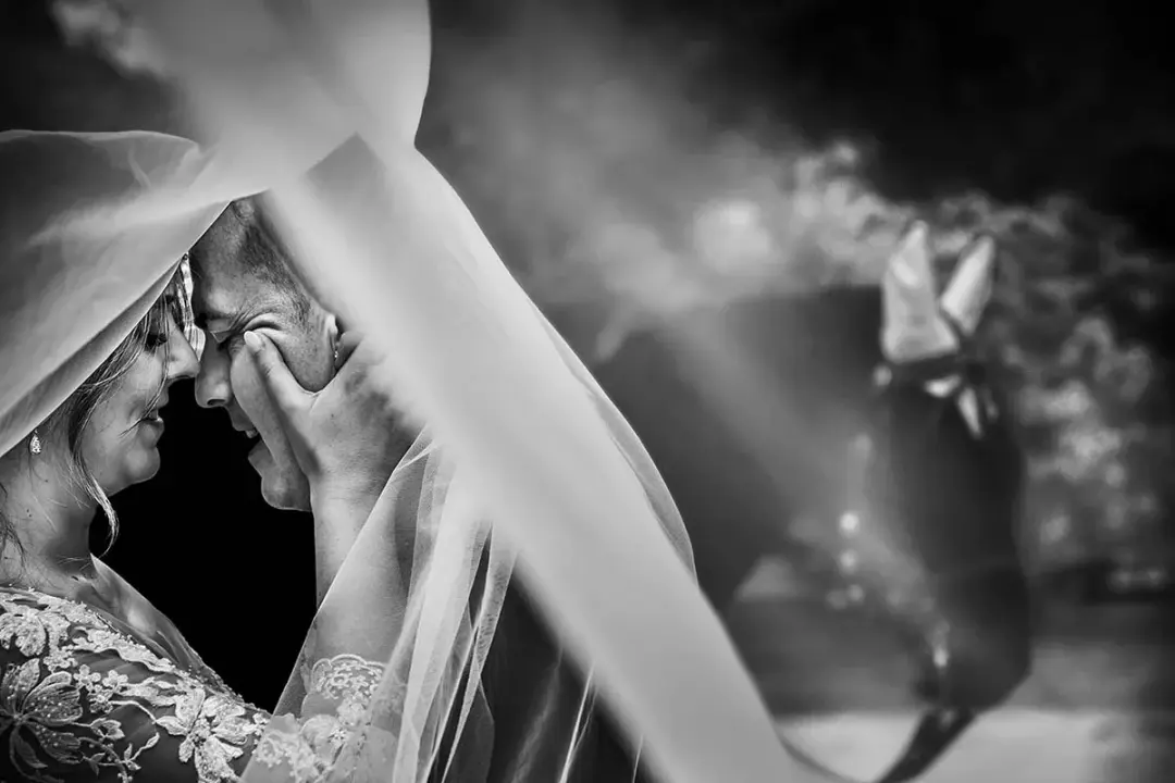 En blanco y negro, la novia y el novio se abrazan casi nariz con nariz bajo un velo translúcido que ella sostiene en alto, con haces de luz filtrándose de fondo.
