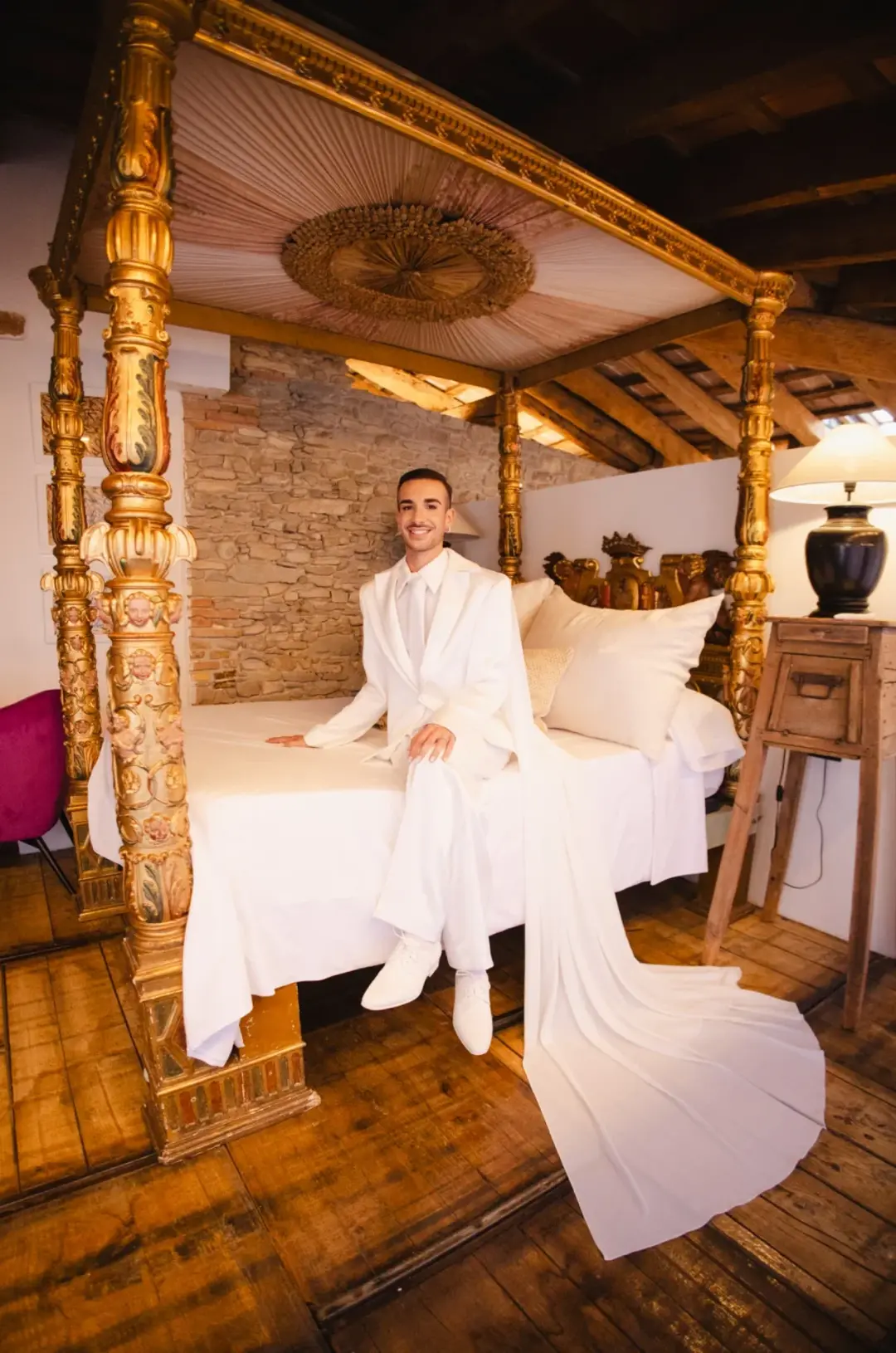 Novio sentado sonriente sobre una cama con dosel dorado tallado, vestido con traje blanco de chaqueta larga tipo capa, en una habitación rústica con paredes de piedra y vigas de madera.