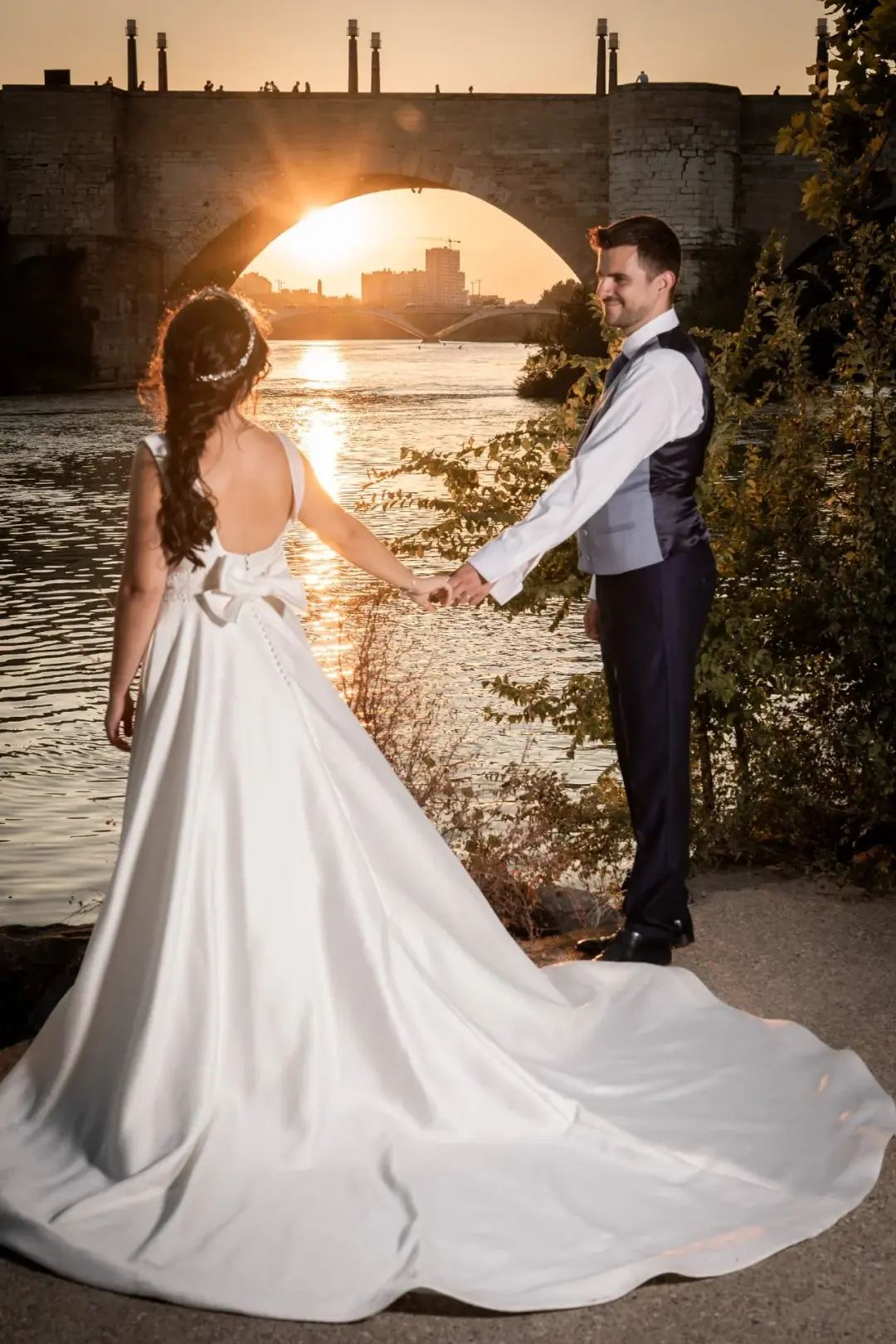 Los novios se dan la mano junto a un río al atardecer, vistos desde atrás de ella con la larga cola del vestido extendida sobre el suelo, mientras el sol se pone justo bajo el arco de un puente de piedra.