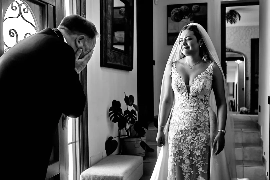 Fotografía en blanco y negro del primer vistazo, con el padre de la novia cubriéndose la cara emocionado y llorando junto a una puerta, mientras ella lo observa con gesto contenido vestida ya de novia con encaje.