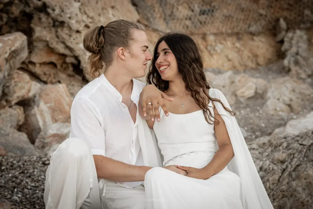 Pareja sentada sobre rocas, mirándose sonrientes, ella con el brazo apoyado en el hombro de él mostrando su anillo de compromiso, ambos vestidos de blanco.