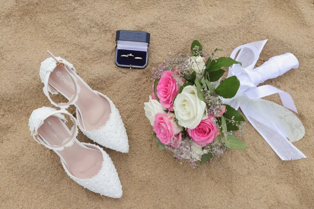 Composición cenital sobre arena de playa con los zapatos de novia de encaje, un estuche abierto con dos anillos, un ramo de rosas rosas y blancas con flor de bebé, y un lazo de raso blanco extendido.
