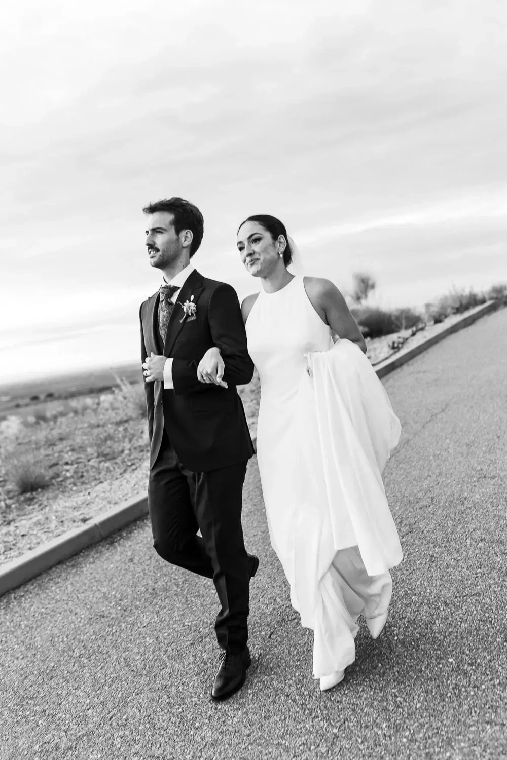 Foto en blanco y negro de los novios caminando del brazo por una carretera solitaria con paisaje árido de fondo, ella sujetando la falda del vestido al viento, ambos con la mirada al frente.