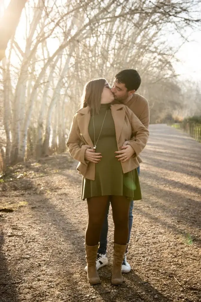 Pareja besándose de pie en un camino de tierra bordeado de árboles sin hojas, a contraluz invernal, ella lleva abrigo camel y vestido verde, él abrigo camel y vaqueros.