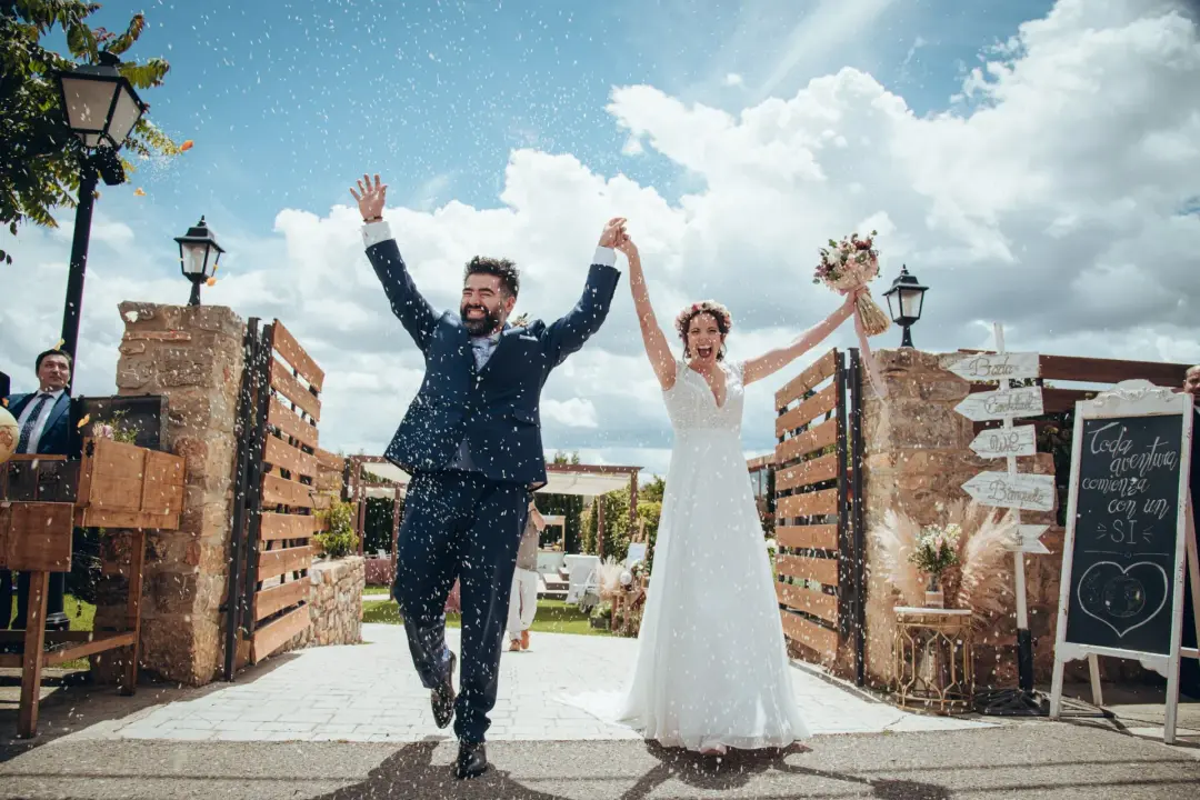 El novio y la novia corren tomados de la mano y riendo bajo una lluvia de arroz, junto a un cartel de madera que dice 'Toda aventura comienza con un sí' y un cielo azul con nubes.
